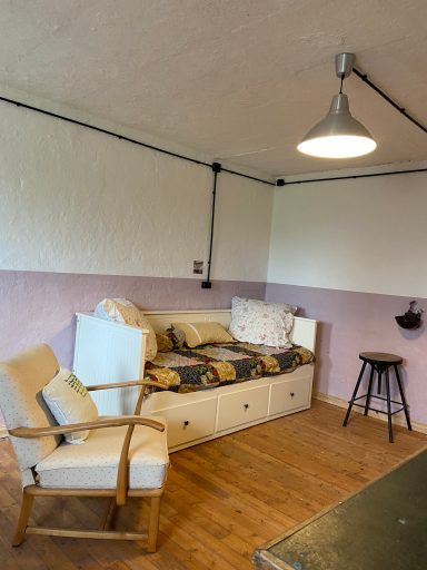 Gemütliche Ecke mit einem Bett, einem Stuhl und einem Hocker in einem schlichten Raum.